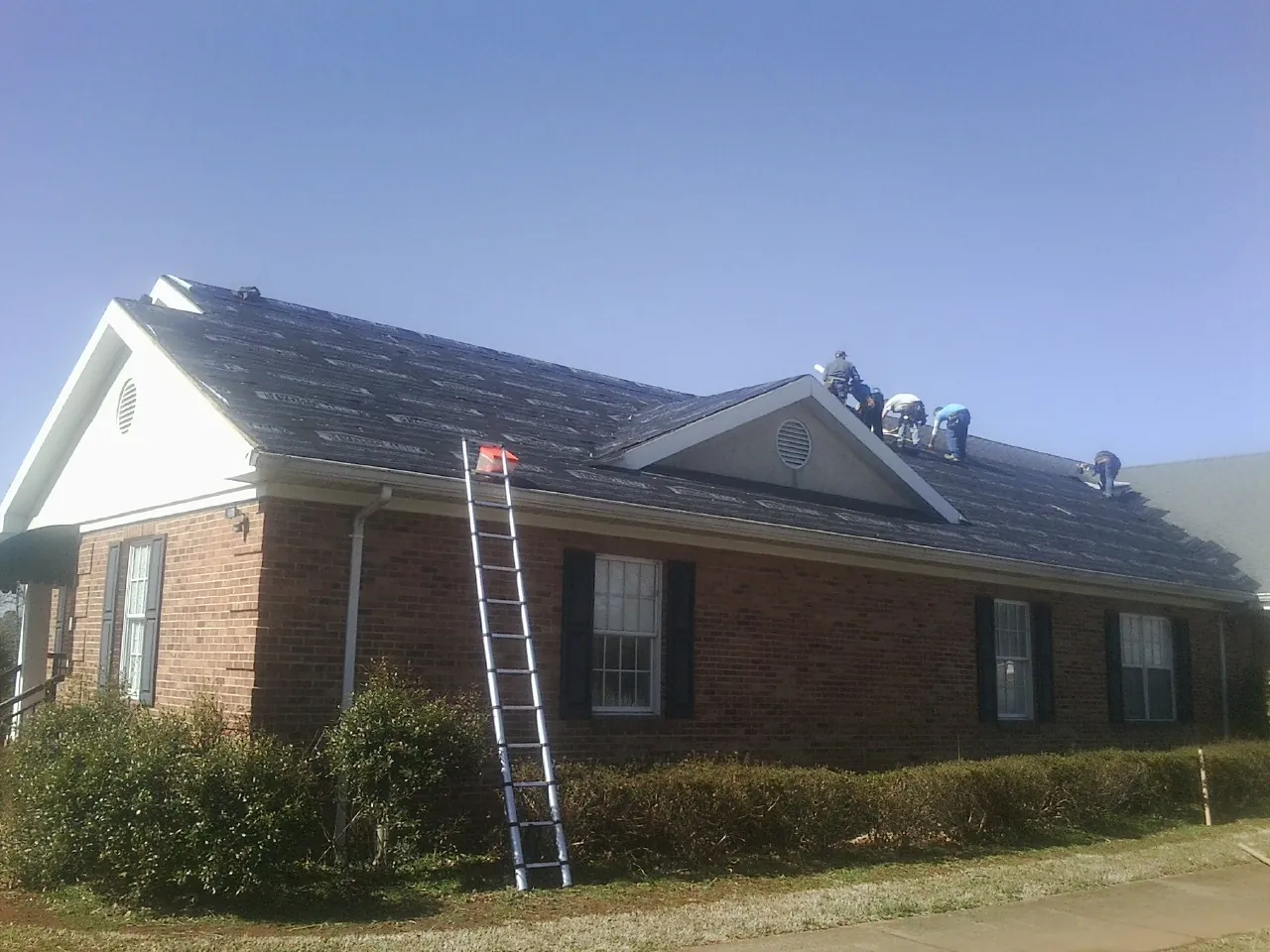 Crew tearing shingles off a brick ranch roof with a ladder set — Metal Roof Repair work by our crew in Windom