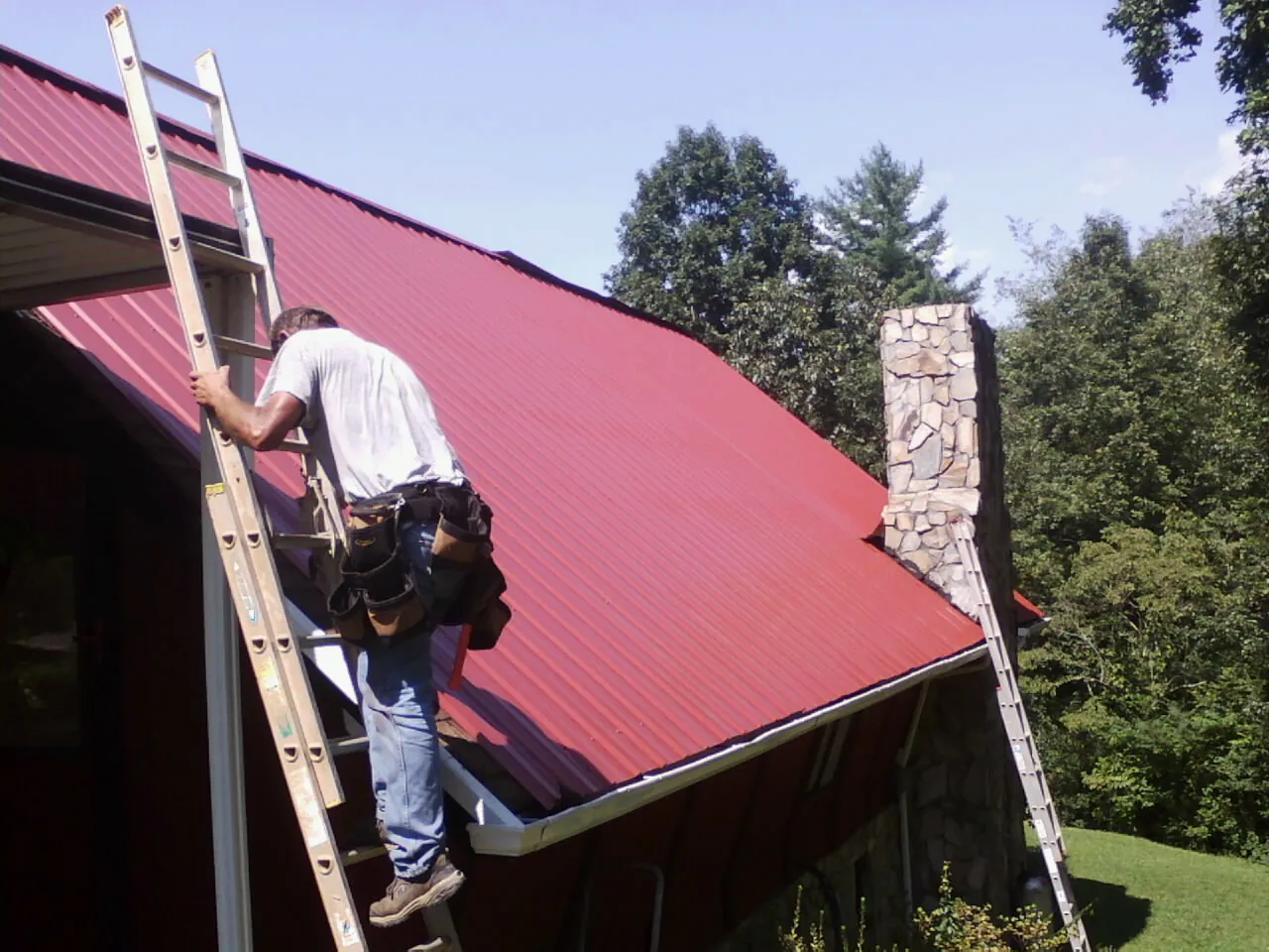 Roofer on a ladder beside a red metal roof and stone chimney — Soffit & Fascia Repair by Roofing Brothers in Windom